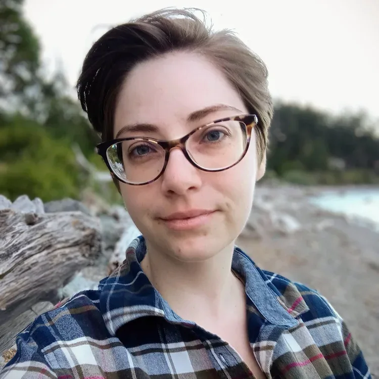Ashley is a white nonbinary person with short brown hair. They are wearing a blue, brown, red, and white plaid shirt and brown mottled glasses. The out of focus shoreline of La Conner WA is in the background.